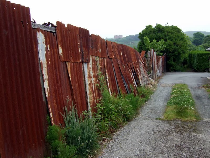 Weathered corrugated steel sheets forming a long boundary fence beside a quiet lane, the ribs and uneven tops showing how a metal fence can offer footholds if it is not detailed as a barrier.