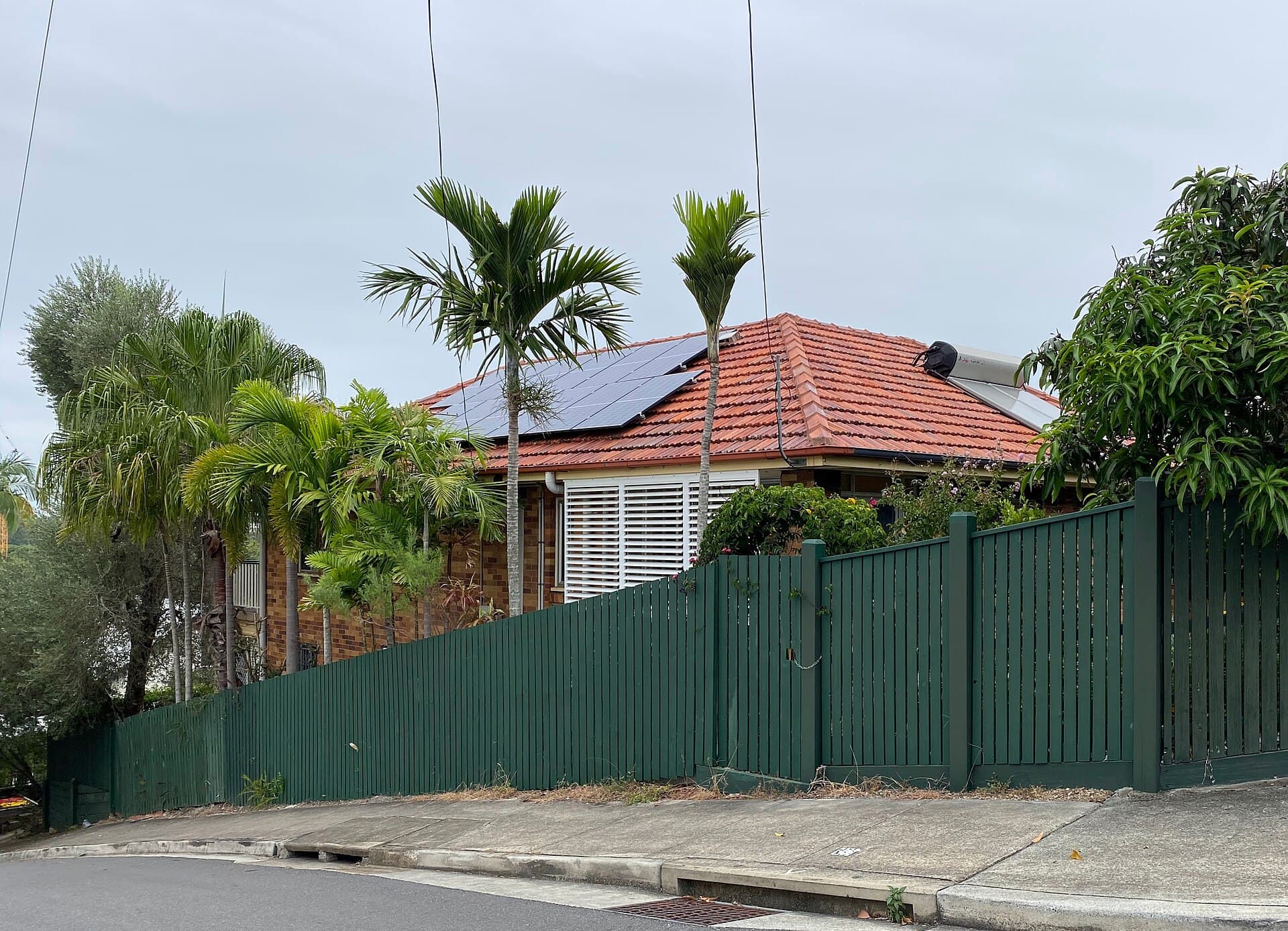 A long green paling fence running the street boundary of a brick Queenslander in West End, Brisbane, the sort of suburban dividing line that is sometimes asked to work as a pool barrier as well.